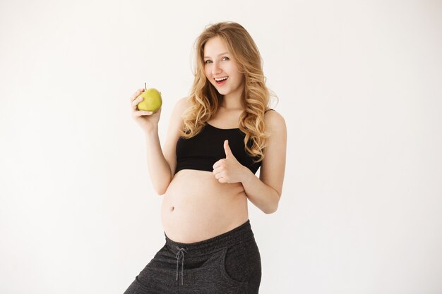 Bela jovem mãe com cabelos loiros em roupas pretas, sorrindo, segurando a maçã perto da barriga de grávida, aparecendo o polegar