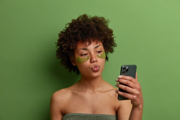 Bela jovem modelo feminina com cabelo afro encaracolado, aplica manchas verdes de hidrogel para reduzir o problema de olheiras, tira selfie no celular, mantém os lábios arredondados, enrolada em uma toalha de banho