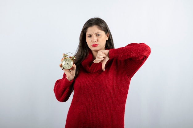 Bela jovem morena com um despertador mostrando o polegar para baixo. Foto de alta qualidade