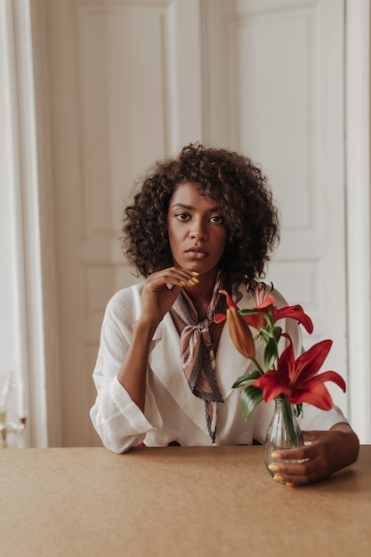 Foto grátis bela jovem morena encaracolada com blusa branca elegante se inclina sobre a mesa de madeira, olha para a frente e toca o vaso com flores vermelhas