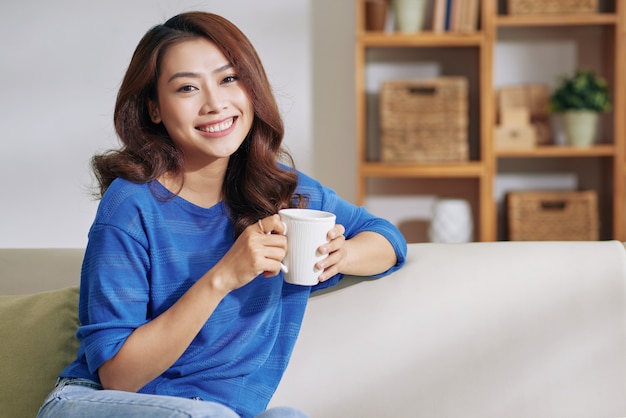 Bela jovem mulher asiática, sentada no sofá em casa com caneca e sorrindo