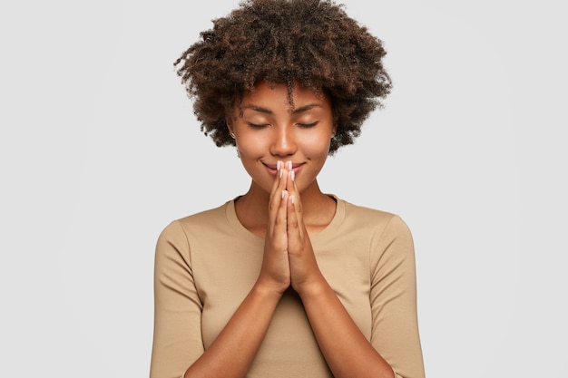Bela jovem negra em pose meditativa, desfruta de uma atmosfera pacífica e dá as mãos em gesto de oração