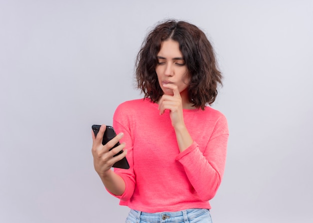 Foto grátis bela jovem pensativa segurando um telefone celular e colocando a mão no queixo em uma parede branca isolada com espaço de cópia