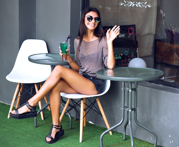 Bela jovem sentada sozinha no café da cidade de rua, olhar hippie minimalista de verão, beber um cocktail e pedir garçom.