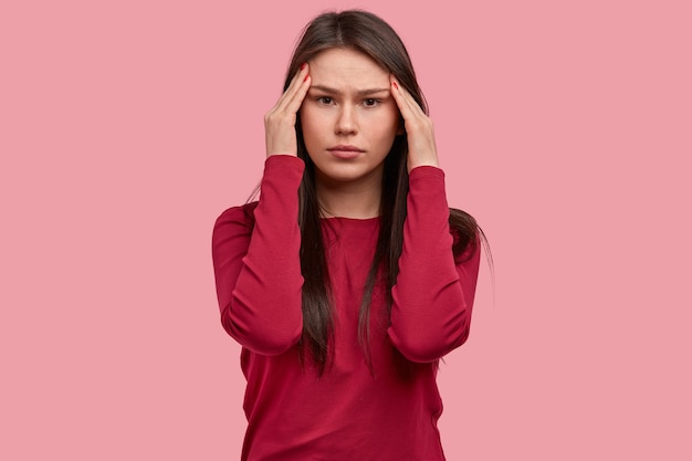 Bela jovem séria segura as têmporas, sente-se pressionada enquanto resolve o quebra-cabeça, usa um suéter vermelho, modelos contra um fundo rosa, está sobrecarregada