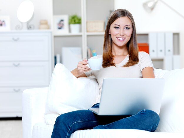 Bela jovem sorridente com laptop e uma xícara de café no sofá