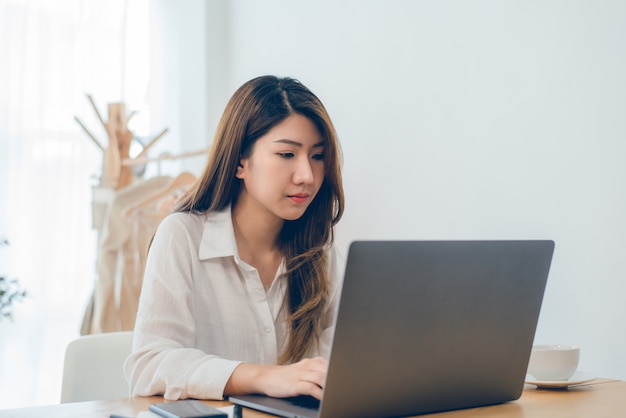 Bela jovem sorridente mulher asiática trabalhando no laptop enquanto em casa no espaço de trabalho de escritório