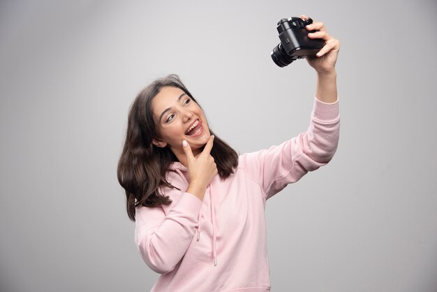 Bela jovem tirando uma selfie com a câmera sobre uma parede cinza.