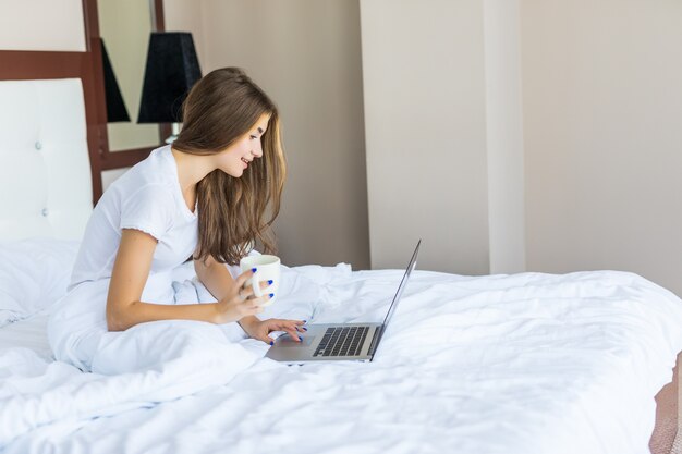 Bela jovem tomando café e navegando na Internet em seu laptop enquanto está sentada na cama e sorrindo para a câmera