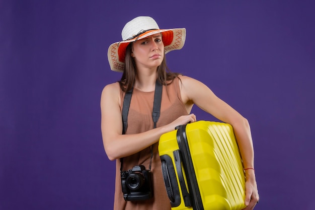 Bela jovem viajante com chapéu de verão com mala amarela e câmera parecendo confusa, sem resposta em pé sobre um fundo roxo