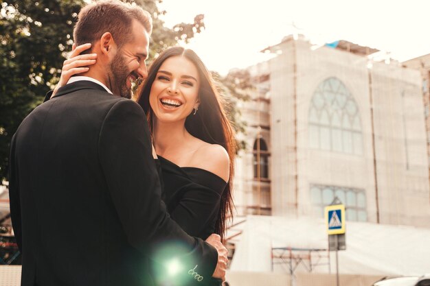 Bela moda mulher e seu namorado bonito de terno Modelo morena sorridente sexy em vestido de noite preto Casal elegante posando na rua ao pôr do sol Homem brutal e sua fêmea ao ar livre