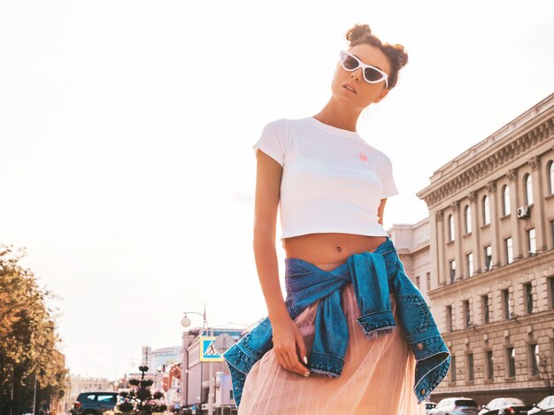 Bela modelo sorridente com penteado de chifres, vestido com roupas de jeans de jaqueta hipster de verão. Garota despreocupada sexy, posando na rua. Mulher na moda engraçada e positiva, se divertindo em óculos de sol