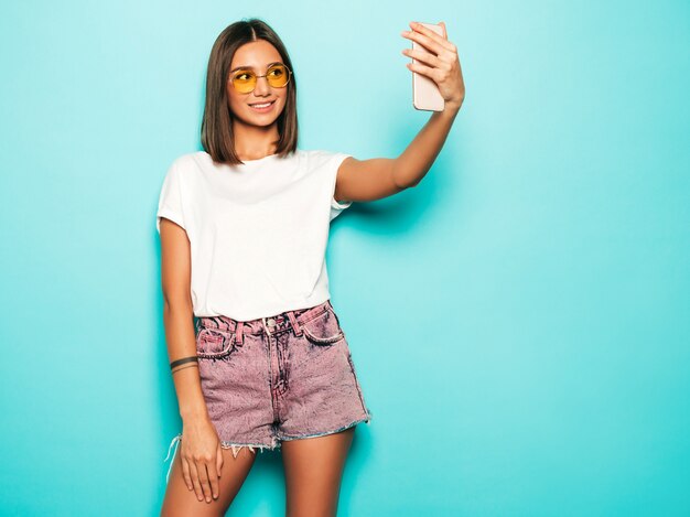 Bela modelo sorridente, vestida com roupas de verão hipster. Garota despreocupada sexy, posando no estúdio perto da parede azul em shorts jeans. Mulher na moda e engraçada, tirando fotos de auto-retrato de selfie no smartphone