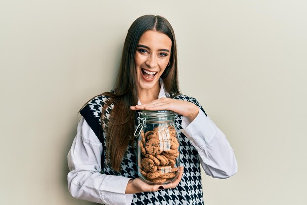Bela morena jovem segurando o pote de biscoitos de chocolate sorrindo e rindo muito em voz alta porque piada louca e engraçada.