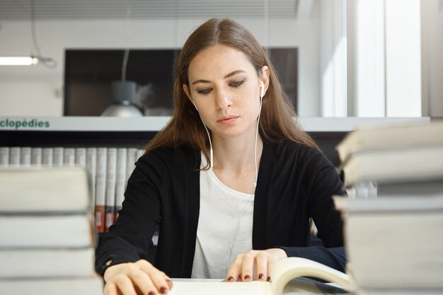 Bela mulher adolescente com cabelos longos morena vestindo uniforme estudando livro ou manual, ouvindo sua música favorita com fones de ouvido enquanto está sentado na biblioteca da escola