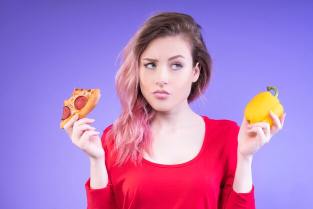 Bela mulher escolhendo entre uma fatia de pizza e um pimentão amarelo