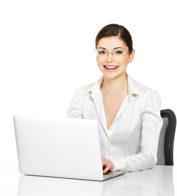 Bela mulher sorridente se senta na mesa com o laptop na camisa branca - isolado no branco.