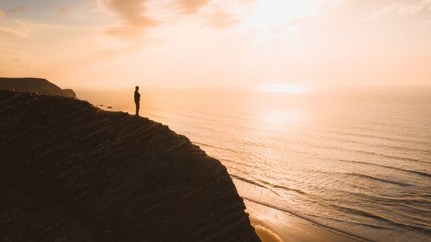 Bela vista de uma pessoa em pé em um penhasco sobre o oceano ao pôr do sol no Algarve, Portugal