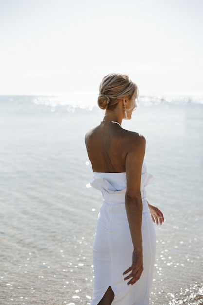 Bela vista traseira de uma jovem loira de vestido branco está andando perto do mar no dia ensolarado