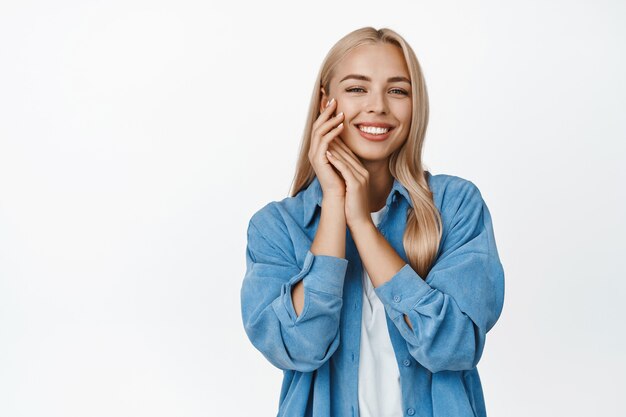 Beleza e bem-estar feminino. Loira tenra de mãos dadas perto da pele macia e brilhante do rosto, olhando e sorrindo, de pé sobre o branco.