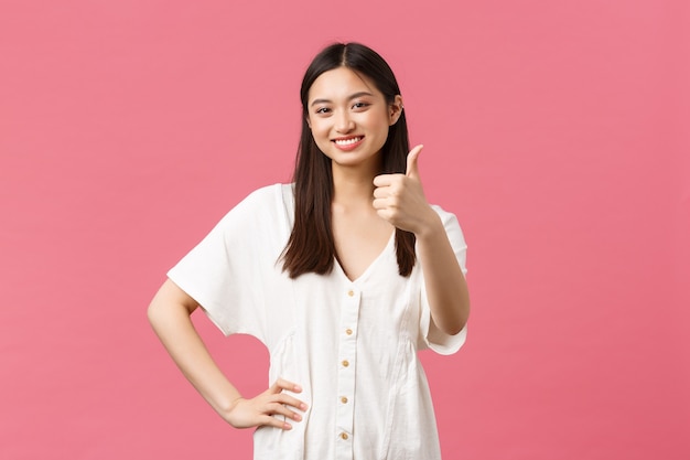 Beleza, emoções das pessoas e conceito de férias e lazer de verão. Menina asiática fofa satisfeita em um vestido branco, mostrando o polegar em aprovação, gosto e concorda, avalie um produto excelente, fundo rosa