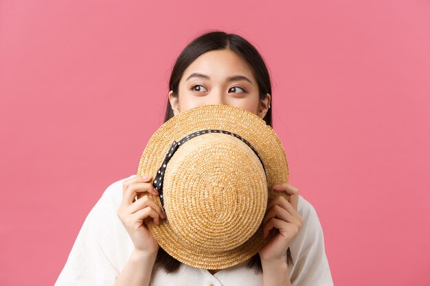 Beleza, emoções das pessoas e conceito de lazer de verão. Close da linda garota asiática sonhadora, escondendo o rosto atrás de um chapéu de palha e espiando para a esquerda para o banner promocional, sorrindo com os olhos, fundo rosa