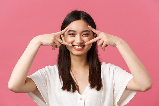 Beleza, emoções das pessoas e conceito de lazer de verão. Close-up de kawaii sorrindo tenra mulher em vestido branco, mostrando o símbolo da paz sobre os olhos e sorrindo com dentes brancos, fundo rosa.