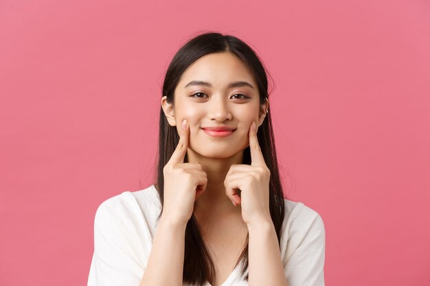 Beleza, emoções das pessoas e conceito de lazer de verão. Close-up de mulher asiática engraçada e bonita com covinhas kawaii, tocando as bochechas e sorrindo feliz, em pé fundo rosa.