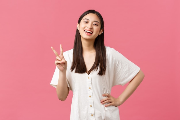 Beleza, emoções das pessoas e conceito de lazer de verão. menina japonesa feliz e entusiasmada rindo e sorrindo, mostrando o símbolo da paz kawaii em um vestido branco fofo, fundo rosa