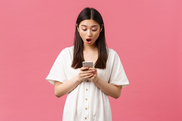 Beleza, emoções das pessoas e conceito de tecnologia. Menina asiática chocada e assustada lendo grandes notícias no celular, olhar para a tela do smartphone espantada com o queixo caído, fundo rosa de pé.