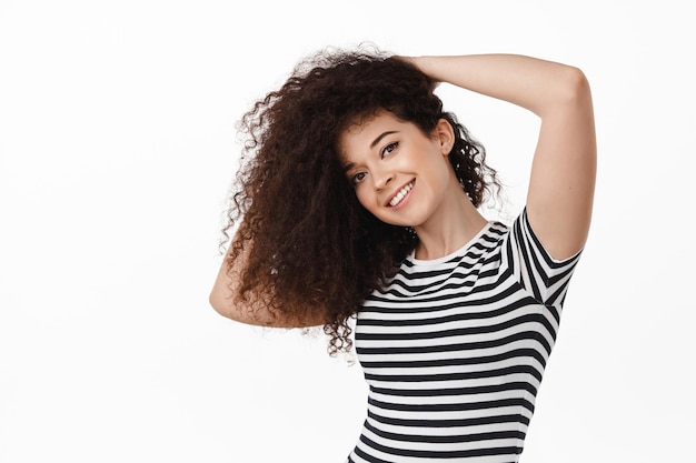 Beleza. jovem morena atraente com cabelo longo e saudável encaracolado, penteado de chicote e sorrindo, cuidando do corte de cabelo, de pé contra um fundo branco