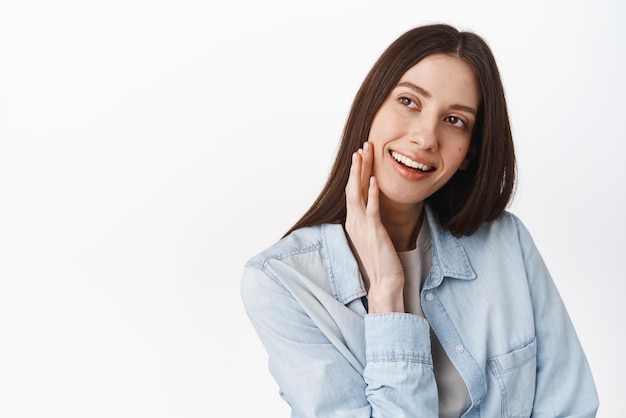 Beleza Menina de beleza adolescente tocando pele fresca perfeita, lisa e limpa, sem maquiagem, rosto nu natural, olhando de lado para o logotipo de texto promocional em pé contra um fundo branco