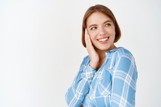 Beleza. Retrato de mulher feliz tocando a bochecha e olhando de lado no espaço vazio. Rapariga com pouca maquilhagem natural, pele saudável e perfeita após cosméticos de cuidados diários, parede branca