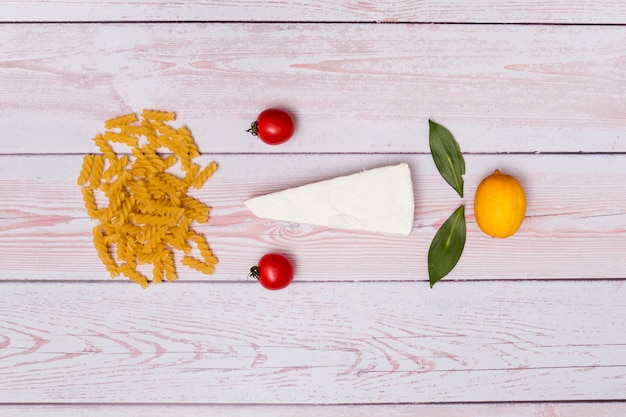 Belo arranjo de macarrão fusilli cru; tomates; queijo; folhas de louro e limão no fundo de madeira