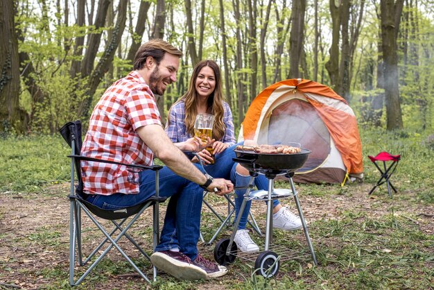 Belo homem feliz preparando churrasco ao ar livre enquanto bebe cerveja