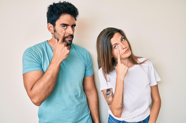 Belo jovem casal de namorado e namorada juntos apontando para o olho observando você gesticular expressão suspeita