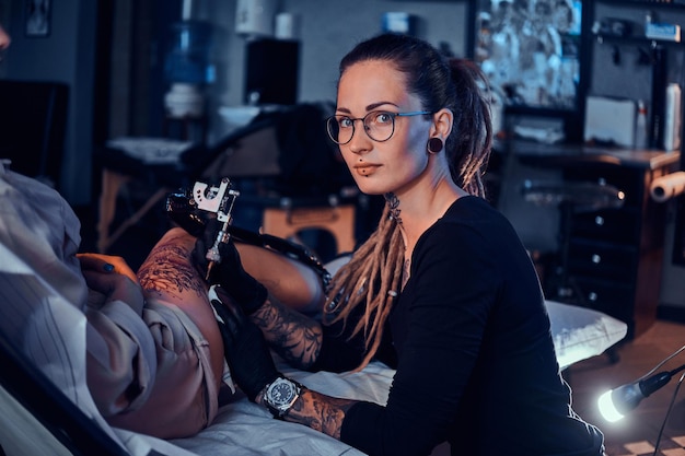 Belo mestre criativo com dreadlocks está trabalhando em uma nova paz de tatuagem de perna grande para o cliente.