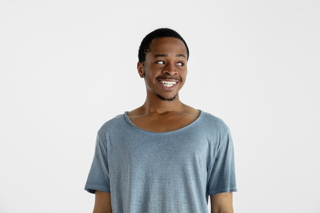 Belo retrato de meio corpo masculino isolado na parede branca. jovem emocional afro-americano de camisa azul. expressão facial, emoções humanas, conceito de anúncio. sorrindo, olhando para o lado.