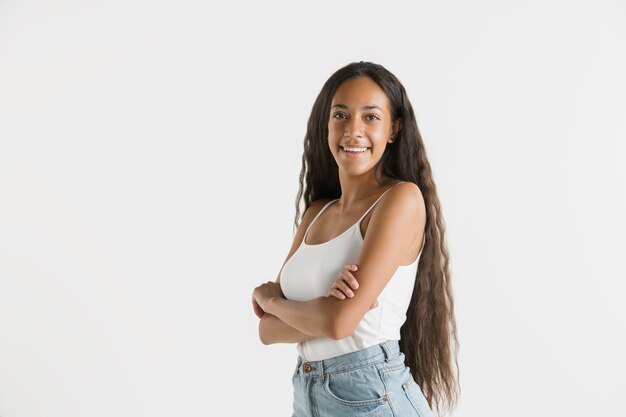 Belo retrato feminino de meio comprimento isolado na parede branca. Jovem emocional afro-americana com cabelo comprido. Expressão facial, conceito de emoções humanas. De pé e sorrindo.