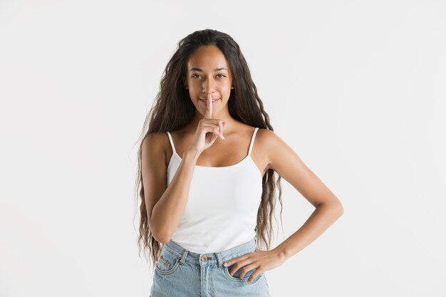 Belo retrato feminino de meio comprimento isolado na parede branca. Jovem emocional afro-americana com cabelo comprido. Expressão facial, conceito de emoções humanas. Sussurrando um segredo.