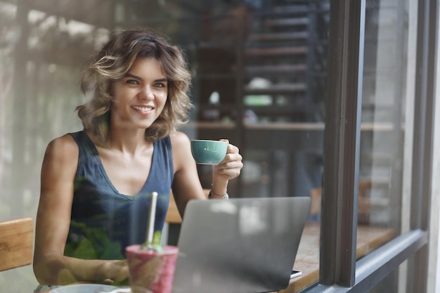 Bem sucedido sortudo atraente Europeu mulher loira cabelo curto encaracolado segurar xícara de café trabalhando laptop editar fotografia freelance sentar bar da janela café olhar fora busca inspiração Nômade digital