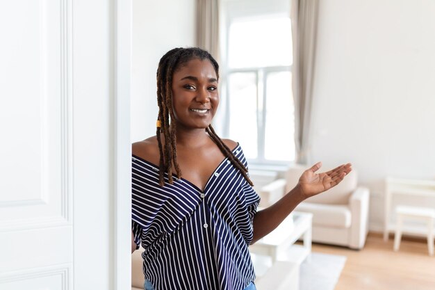 Bem-vindo Retrato de alegre mulher africana convidando o visitante a entrar em sua casa jovem feliz em pé na porta do apartamento moderno mostrando a sala de estar com a mão