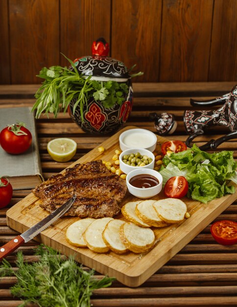 Bife com batatas assadas redondas em uma mesa de madeira, vista lateral, com salada verde, feijão e maionese