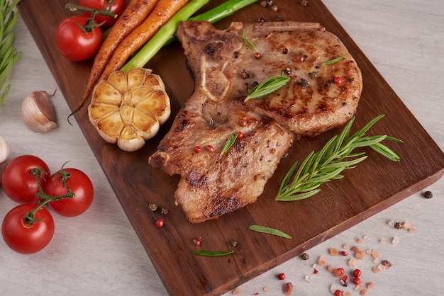 Bife de porco grelhado de um churrasco de verão servido com legumes, aspargos, cenouras, tomates frescos e especiarias. Bife grelhado na tábua de madeira na superfície da pedra. vista do topo.