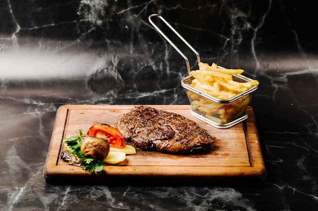 Bife do lombo com legumes grelhados e batatas fritas em uma placa de madeira.