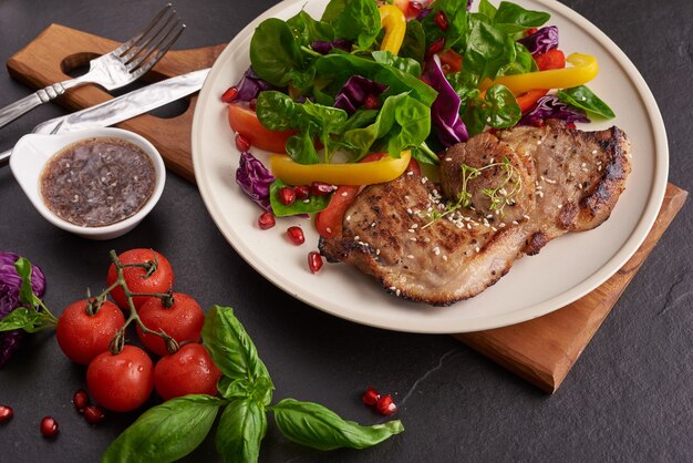 Bife grelhado com vegetais frescos, pimentão, tomate, cebola roxa, pimenta rosa e especiarias. comida saborosa feita em casa. conceito de refeição saborosa e saudável. superfície de pedra preta. bife de porco com salada