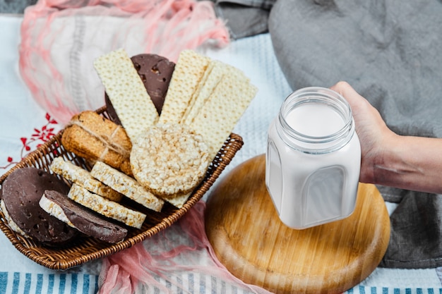 Biscoitos em uma cesta e mão segurando um pote de leite sobre uma toalha de mesa.