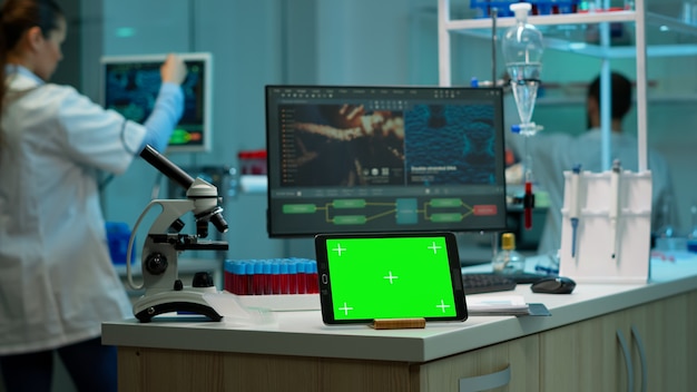 Bloco de notas com tela verde trabalhando em laboratório com monitor de simulação, display de chroma key, enquanto engenheiro profissional testa a evolução do vírus em segundo plano. Laboratório de desenvolvimento de alta tecnologia.