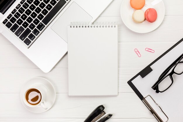 Bloco de notas em espiral com laptop, biscoitos, xícara de café com prancheta e óculos na mesa de escritório branco