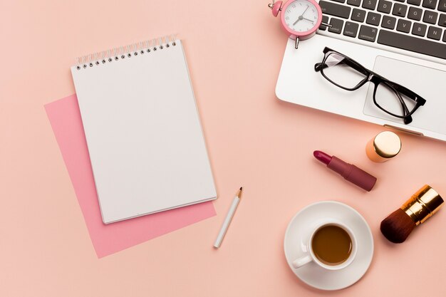 Bloco de notas em espiral com laptop, óculos, laptop, produto de maquiagem e xícara de café em fundo colorido
